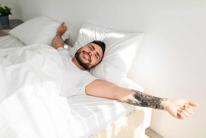 Comfortable sleep. Wellslept tattooed man waking up in morning and stretching hands, lying in bed
