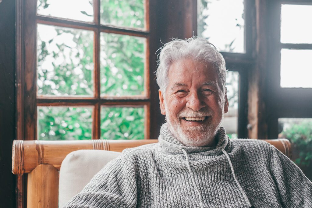 An elderly man smiling at the camera while sitting comfortably in his home.